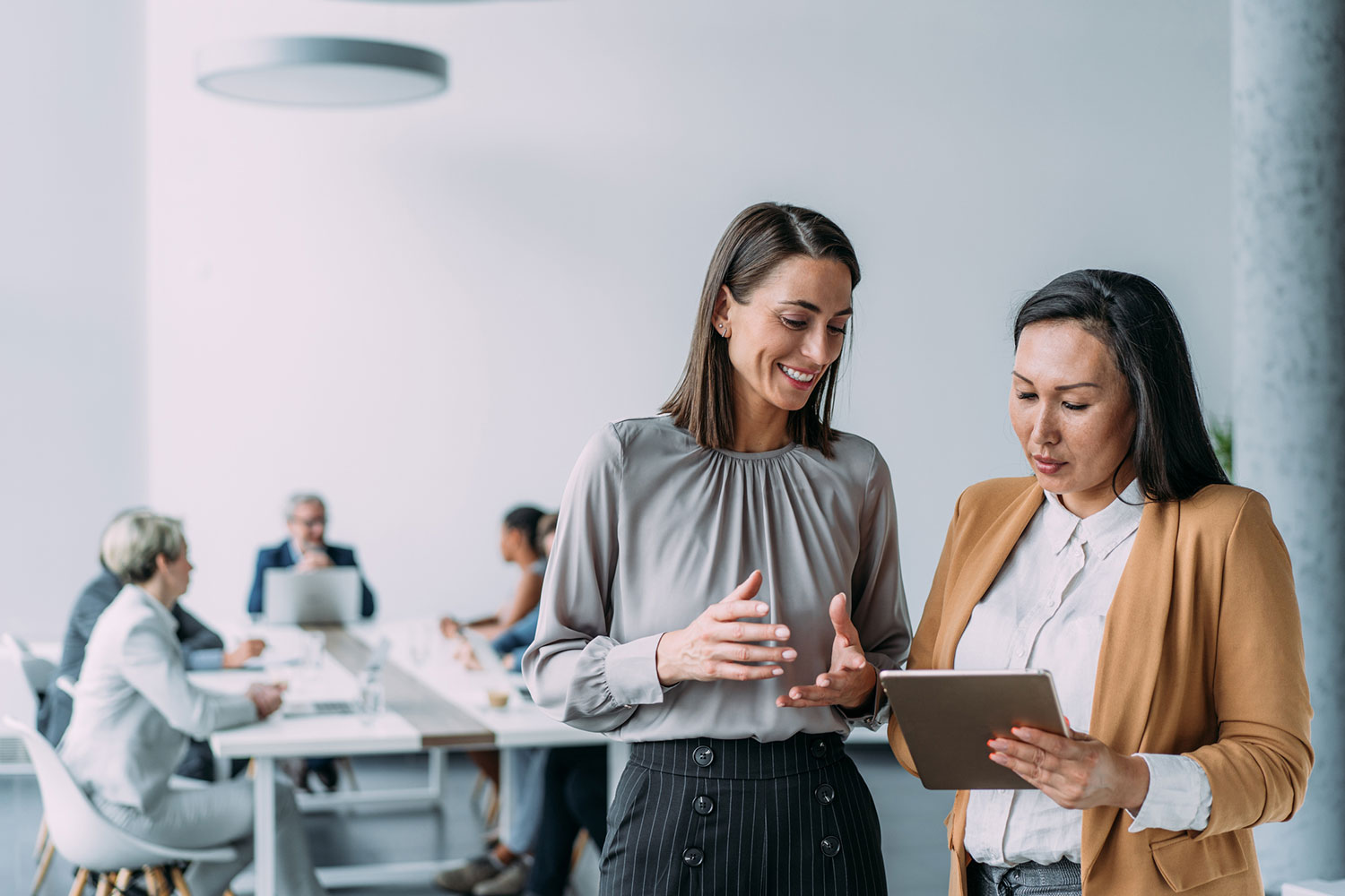 Image of two business women standing talking next to a board room table, one holds a tablet.