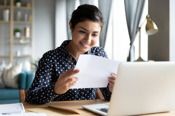 Smiling young woman sitting at a desk with laptop, looking at papers